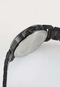 Montre en acier inoxydable noir avec un boîtier ronde, bracelet en métal et une couronne proéminente. Présente un fond en argent avec une gravure "HUGO".