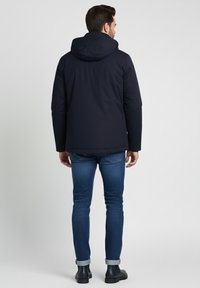 Hombre vestido con una chaqueta con capucha en color azul marino, jeans azules enrollados y botas oscuras, de pie de espaldas contra un fondo claro y liso.