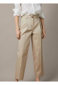 Pantalon beige sur mesure avec une coupe droite, des plis avant et une fermeture par bouton. Le tissu a une texture lisse avec une finition soignée.