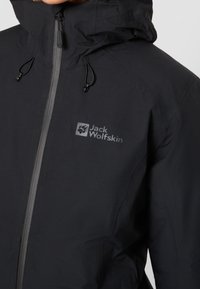 Sort vandtæt jakke med lynlåsgardin, justerbar snøre i huen og Jack Wolfskin-logoet. Materialet har en glat overflade.