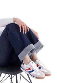 Zapatillas blancas con acentos en rojo y azul, de material de ante y malla, con cordones blancos y suela de goma. Combinadas con pantalones de mezclilla oscuros y acampanados.