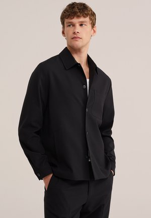 Fiatal férfi göndör hajjal, fekete gombos dzsekit visel fehér ing felett, kezét zsebében tartva, oldalra néz egy egyszerű háttér előtt.