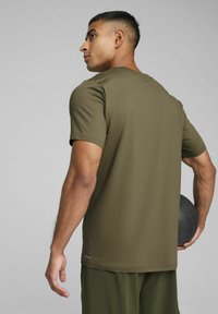 Camiseta deportiva verde oliva hecha de tela suave, de manga corta, con cuello redondo y un pequeño logo en el dobladillo inferior, usada con ropa de entrenamiento.