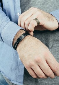 Bracciale in pelle intrecciata nera con chiusura rettangolare in argento. Un anello in bronzo testa di moro con texture è visibile su un dito. Camicia casual azzurro chiaro a maniche lunghe in evidenza.
