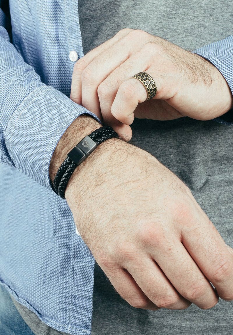 Bracciale in pelle intrecciata nera con chiusura rettangolare in argento. Un anello in bronzo testa di moro con texture è visibile su un dito. Camicia casual azzurro chiaro a maniche lunghe in evidenza.