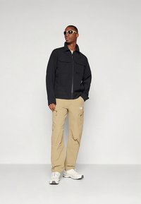Black zip-up jacket with two chest pockets, paired with beige pants. The outfit is completed with white sneakers and sunglasses.