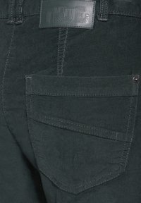Gros plan sur la poche arrière d'un pantalon en velours côtelé vert foncé avec des détails cousus et une étiquette de marque en cuir "TRUE SELF" au-dessus de la poche.