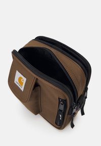 Borsa di stoccaggio in tessuto marrone con due scomparti zip, dotata di tasca frontale e una toppa con logo Carhartt. Superficie texture e zip robuste.