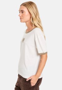 Witte T-shirt met korte mouwen en olijfgroen accent, voorzien van een rechthoekig logo op de borst. Zachte stof met een relaxed fit.