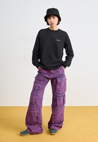 Chemise noire à manches longues avec logo, pantalon cargo violet ample avec plusieurs poches, et chaussures vertes sur un sol jaune.