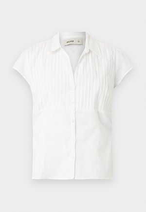 Bijela košulja kratkih rukava s gumbima sprijeda, s okomitim pinprugama na gornjem dijelu i glatkim donjim dijelom. Pamuk.