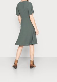 Vihreä lyhythihainen mekko, jossa on levenevä helma, valmistettu sileästä kankaasta. Yhdistettynä ruskeisiin nilkkureihin, mikä tuo esiin yksinkertaisen ja tyylikkään muotoilun.