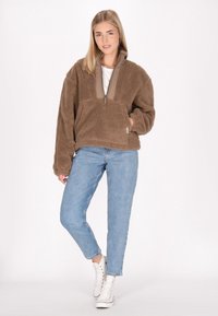 Pullover di pile marrone con zip a metà, dotato di un pannello del collo beige e una tasca frontale, abbinato a jeans azzurri chiari e sneakers bianche.