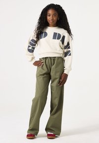 Crèmekleurige sweatshirt met zwarte grafische tekst, gecombineerd met olijfgroene wijde broek. Model draagt bordeauxrode en witte schoenen. Textuurhaar.