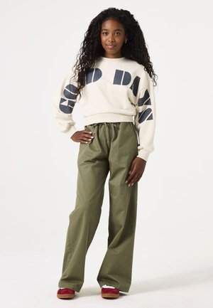 Crèmekleurige sweatshirt met zwarte grafische tekst, gecombineerd met olijfgroene wijde broek. Model draagt bordeauxrode en witte schoenen. Textuurhaar.