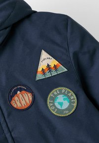 Jūras spilventiņš ar trim auduma plāksteriem: trīsstūra plāksteris ar mežu dizainu, apaļais plāksteris ar vārdu "BRAVE" un plāksteris ar vārdiem "LOVE THE PLANET" un Zemes zīmējumu.