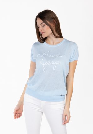 Femme aux cheveux bruns portant un haut en tricot bleu clair avec le texte « Ne jamais abandonner » et un pantalon blanc, debout devant un fond blanc.