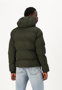 Grønne pufferjakke med hætte, der har en kviltet tekstur, ribbede manchetter og et cropped design. Bæret over lyseblå jeans.