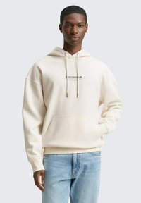 Hoodie beige clair en tissu doux avec une poche avant, un capuchon avec cordon de serrage et un logo imprimé. Assorti avec un jean bleu clair.