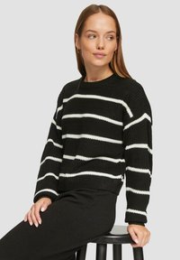Schwarzer Strickpullover mit weißen horizontalen Streifen, gerippten Bündchen und Rundhalsausschnitt. Getragen mit schwarzen Hosen, sitzend auf einem Hocker.
