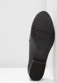 Semelle d'une chaussure en cuir noire montrant un motif de grip texturé avec la marque répétée "Tamaris" et la taille 38 indiquée, sur fond blanc.