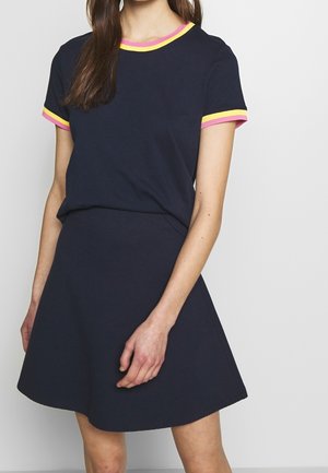 Vrouw draagt een marineblauw T-shirt met korte mouwen met roze en gele accenten en een bijpassende marineblauwe A-lijn rok tegen een effen achtergrond.