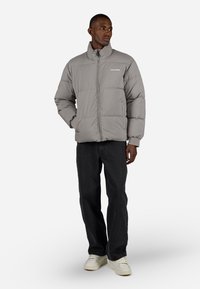 Grå puffjakke med høy krage, glidelås og ribbede mansjetter. Logo trykt på brystet. Kombinert med svarte jeans og hvite joggesko.