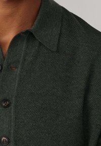 Tamnozeleni pleteni pulover s ovratnikom, izrađen od teksturirane tkanine s crnim gumbima. Ima mali logo na gumbu.