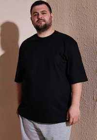 Černé bavlněné tričko s krátkým rukávem, volným střihem a kulatým výstřihem, v kombinaci se šedými tepláky. Texturované světle hnědé pozadí zdi.