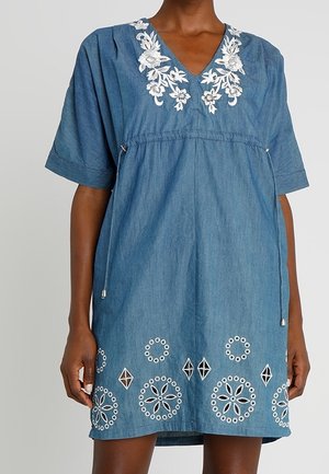 Robe en denim bleu avec broderie florale blanche sur le décolleté et motifs découpés en cercles près de l'ourlet, portée par une femme à la peau foncée.