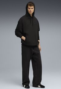 Jonge man met handen in de zak, gekleed in een zwarte hoodie, brede zwarte spijkerbroek en zwarte Puma slippers op een grijze achtergrond.