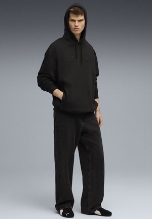 Giovane uomo con le mani in tasca, che indossa un felpa nera con cappuccio, pantaloni larghi neri e pantofole Puma nere su sfondo grigio.