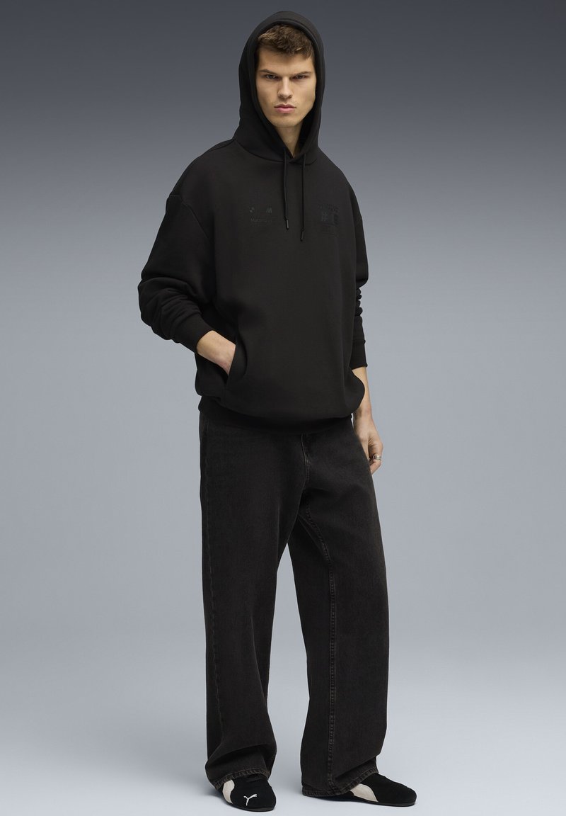 Jonge man met handen in de zak, gekleed in een zwarte hoodie, brede zwarte spijkerbroek en zwarte Puma slippers op een grijze achtergrond.