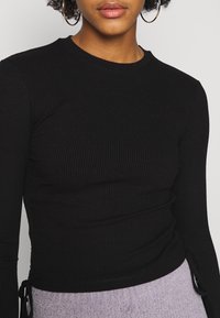Svart ribbad långärmad topp med rund halsringning och knytningar på sidorna, tillverkad av ett stretchigt material för en figurnära passform.