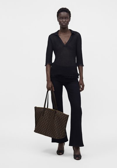 Femme debout portant un haut noir à col en V, un pantalon noir et des talons ouverts noirs, tenant un grand sac fourre-tout marron à motifs par ses poignées.
