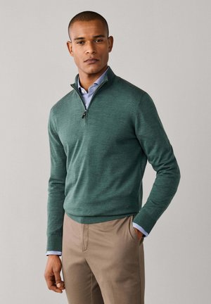Hombre con jersey verde con cremallera hasta el cuarto, sobre camisa azul claro, con pantalones beige, de pie frente a un fondo gris liso.