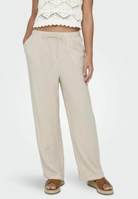 Pantalon large couleur beige clair avec taille à cordon et poches latérales, en tissu texturé. Porté avec un crop top en crochet blanc et des sandales.