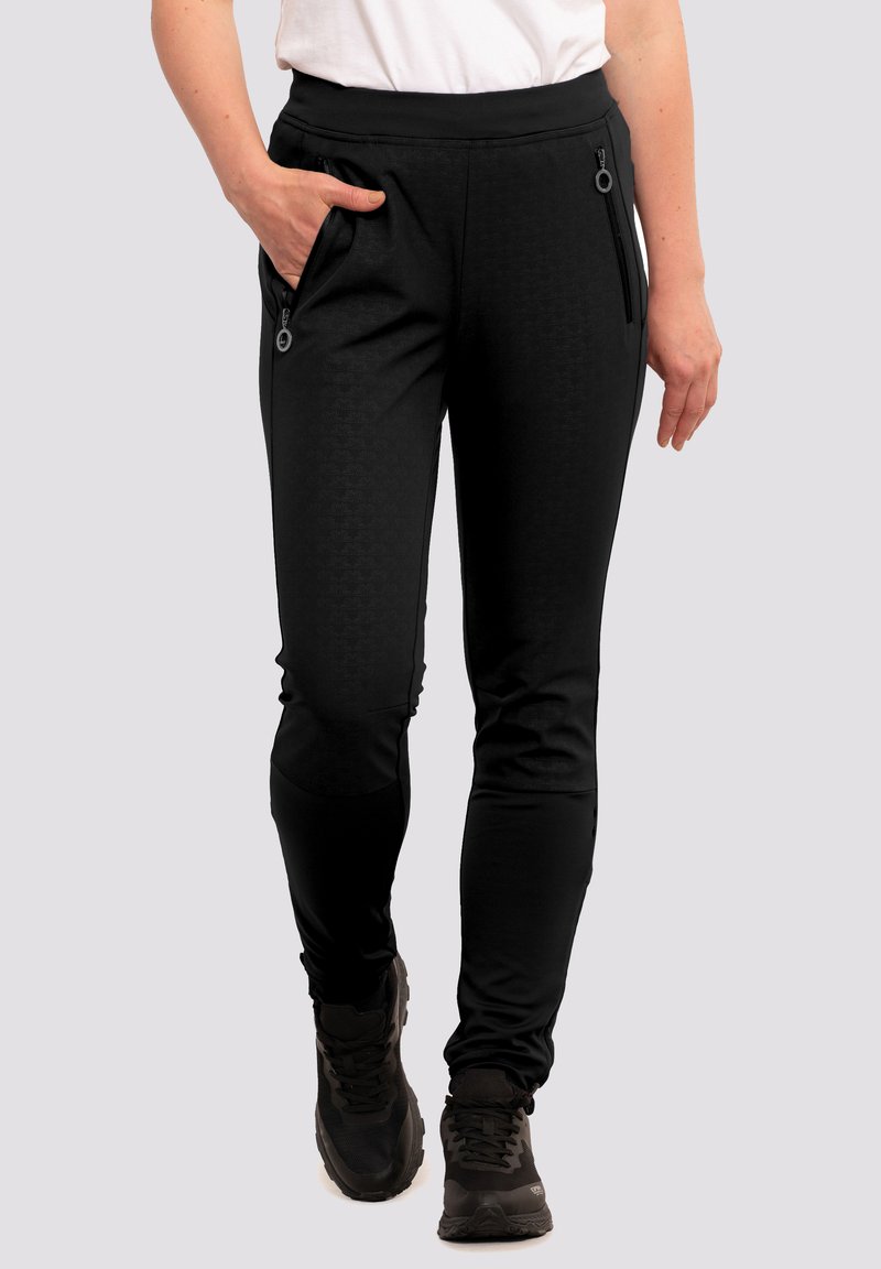 Femme portant un pantalon slim noir avec des poches zippées et des chaussures de sport noires, se tenant avec une main dans sa poche.