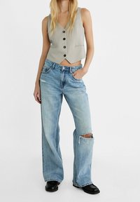 Grå ärmlös väst med V-ringning och knappfront, i kombination med ljusblå vida jeans med rivna detaljer. Svarta skor.