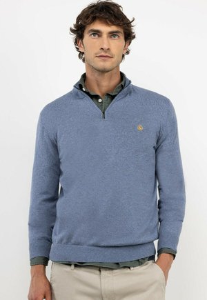 Hombre con un suéter azul con cremallera hasta un cuarto sobre una camisa verde con botones y pantalones beige, de pie frente a un fondo blanco.