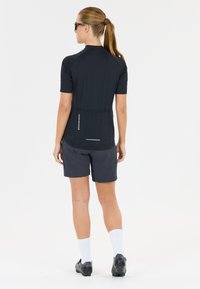 Kvinna i svart cykeltröja och shorts, vita strumpor och svarta cykelskor, stående med ryggen mot en vit bakgrund.