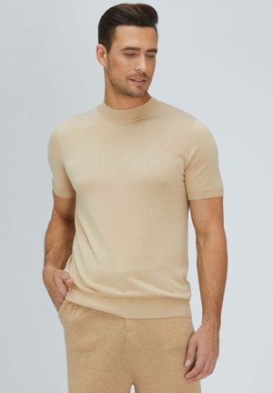 Mann trägt beigen kurzärmeligen Pullover und passende Hose, steht mit einer Hand in der Tasche, schaut leicht nach unten und zur Seite.