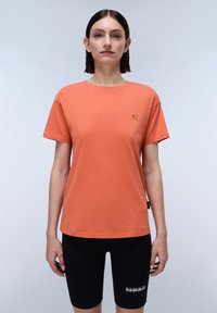 Kurzes oranges T-Shirt aus Baumwolle mit einem kleinen Logo auf der Brust. Kombiniert mit schwarzen Radshorts, der Stoff wirkt glatt.