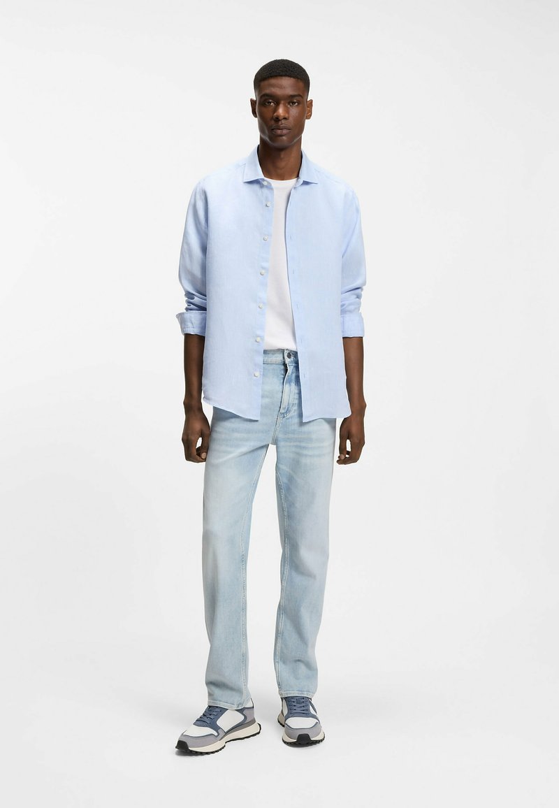 Jeune homme portant une chemise bleu clair ouverte sur un t-shirt blanc, un jean délavé clair et des baskets grises, debout devant un fond blanc.