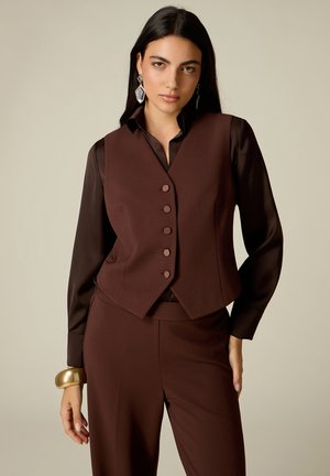 Donna con lunghi capelli scuri indossa un gilet marrone abbottonato sopra una camicia marrone scuro e pantaloni marroni abbinati, accessoriati con grandi orecchini e braccialetto.