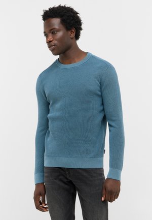 Blauer gestrickter Pullover mit Rundhalsausschnitt, strukturiertem Stoff und gerippten Bündchen. Kombiniert mit dunklen Jeans, was einen lässigen Look erzeugt.