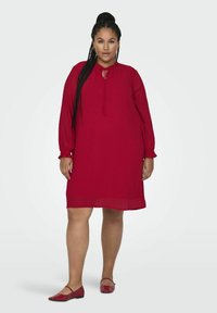 Czerwona sukienka z długimi rękawami, teksturowana tkanina i detal z regulowanym dekoltem. Noszona z dopasowanymi czerwonymi balerinami, na jasnym tle.