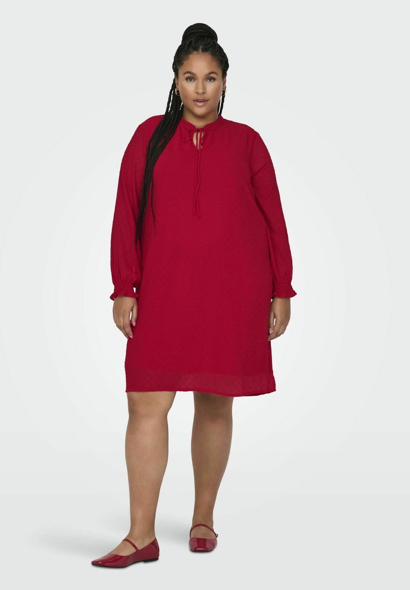 Czerwona sukienka z długimi rękawami, teksturowana tkanina i detal z regulowanym dekoltem. Noszona z dopasowanymi czerwonymi balerinami, na jasnym tle.