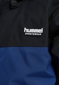 Zwart-blauwe sportjas met een patch met het logo "hummel SPORTSWEAR" op de borst en een zwarte knoop nabij de kraag.