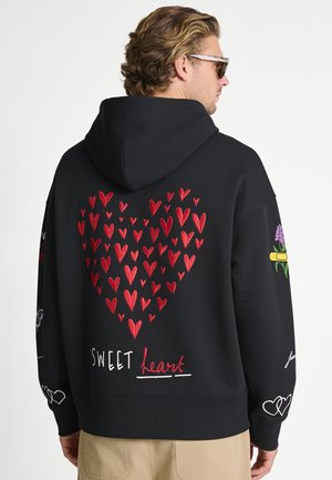 Man wearing black hoodie with red heart pattern, "sweet heart" text on back, floral and heart designs on sleeves, beige pants, and sunglasses.